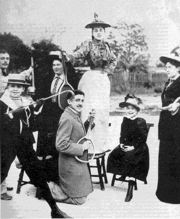 A black and white picture of Marcel Proust, in a grey coat, playing a tennis racket like an air guitar. He's surrounded by ladies wearing hats and long dark dresses.